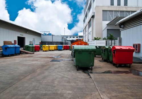 An industrial outdoor area featuring a range of large, wheeled waste bins arranged in a line along a concrete surface. The bins are made of durable plastic with textured surfaces, in colours including blue, green, yellow, and red, indicating different waste types. Behind the bins, there are warehouse-style buildings with metal sheeting exteriors, some with open doors, suggesting storage or operational facilities. To the right, a larger orange waste container is partially visible. The scene is set against a partially cloudy sky, with sunlight illuminating the area and casting soft shadows. The location appears to be part of a commercial waste management site, with the bins and environment consistent with rubbish collection and disposal activities. The setting implies proximity to business premises, possibly in Barkingside, with Commercial Waste Barkingside offering rubbish removal services for local commercial premises.