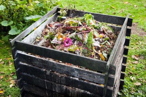 A black, rectangular compost bin made of plastic slats, filled with decomposing organic waste including cut grass, leaves, vegetable peelings, and garden flowers, located outdoors on a grassy area with soil and greenery visible in the background. The compost bin is situated on a garden patio or lawn in Barkingside, with some fallen leaves scattered around its base, reflecting typical rubbish disposal practices related to garden waste management in the local area. The materials inside show various textures such as leafy greens, soft flower petals, and coarse plant debris, contributing to the composting process. The environment is well-lit, suggesting daylight, and the scene conveys a natural setting suitable for waste collection or organic waste disposal services provided by Commercial Waste Barkingside.
