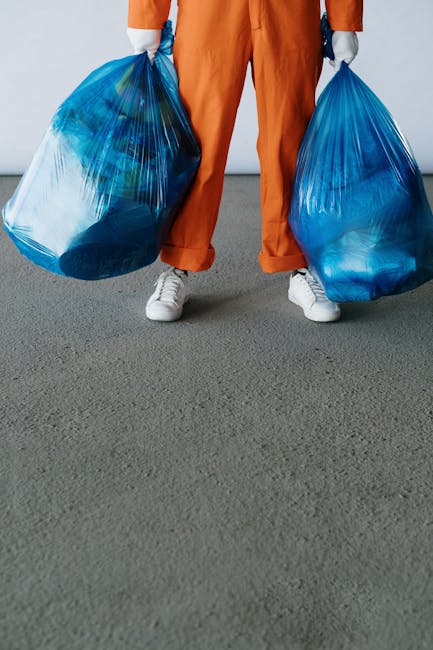 A person dressed in high-visibility orange and yellow safety clothing, including an orange cap, is viewed from above as they collect litter from the edge of a pavement and street. They are holding a g